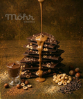 Chocolate stack with hazelnuts and chocolate sauce being poured, on a textured dark background.